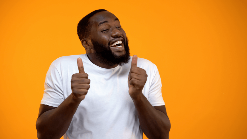 A smiling man in a white shirt gives two thumbs up against a bright orange background
