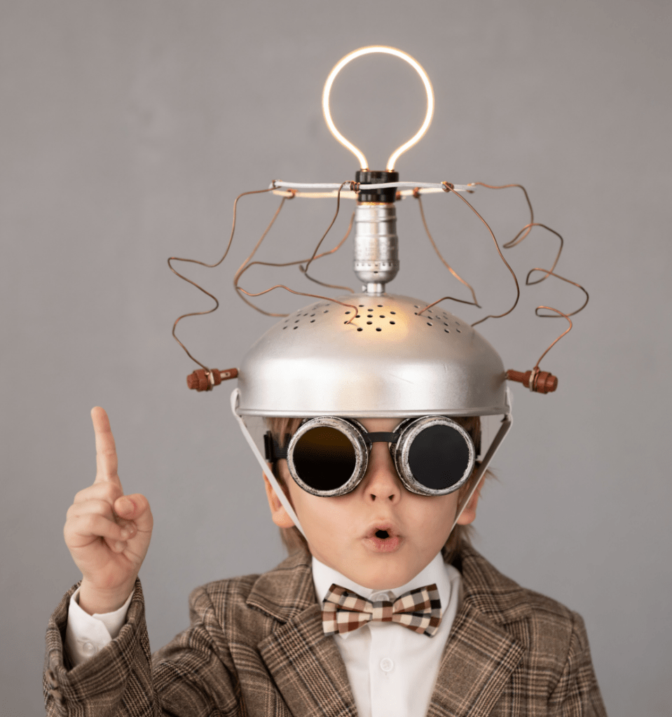 Young boy in a quirky scientist costume, wearing a silver colander hat with wires and goggles, raises a finger, expressing a creative idea.