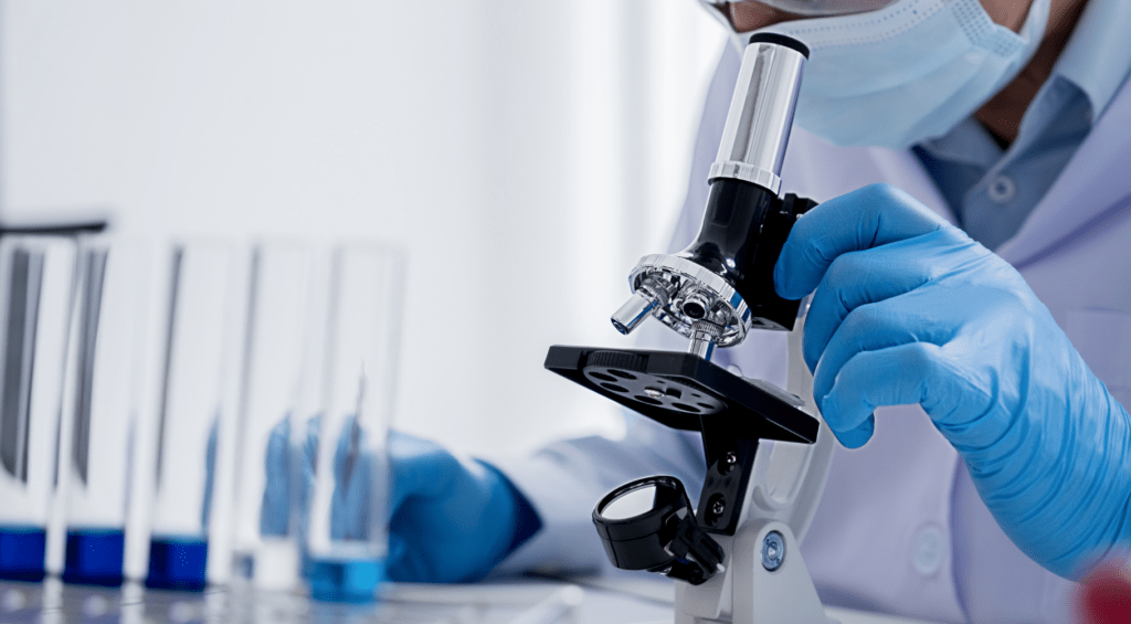 A scientist in protective gear uses a microscope in a lab, surrounded by test tubes filled with blue liquid, conveying focus and precision.